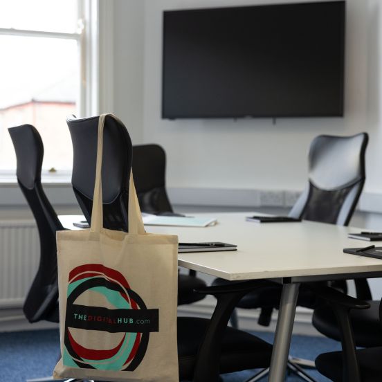 A meeting room with white walls and a blue carpet. There is a large window making the room bright and in the middle of the room is a table with 6 black chairs. There are notebooks and pens on the table and a tote bag hangs from the back of one of the chairs. The TV screen on the wall is turned off.