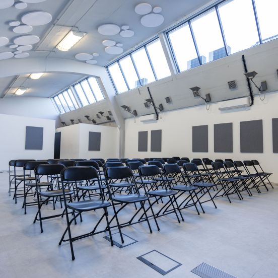 A large bright room painted light grey and white. There are roof lights making the room bright and black chairs laid our in rows ready for an event or performance.