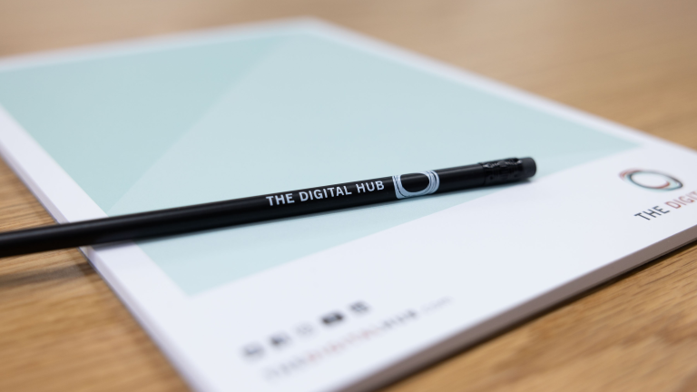 A close up photograph of a writing pad on a wooden desk. A black pencil which is branded with 'The Digital Hub' and its logo is laid upon the paper.