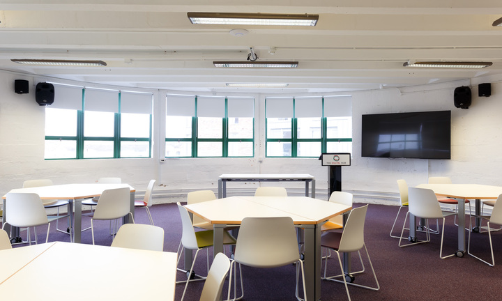 A classroom style room with white round tables and white chairs at each table. There is purple carpet and a tv on the wall.