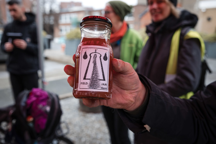 A hand holds a glass jar of golden coloured pear jam. The jar label says 'Field Jam' and has a pencil drawing of a tree against an outline St. Patrick's Tower on it.