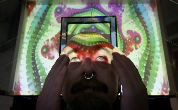 An image of a person with his hands on his head. He is staring upwards as the reflection of a colourful projector cascades over his face.
