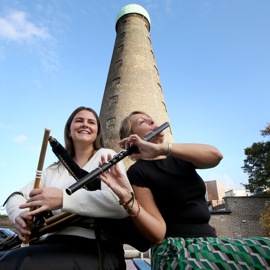 Two people play bagpipes and a flute outdoors in front of a tall brick tower, otherwise known as St Patrick's Tower.